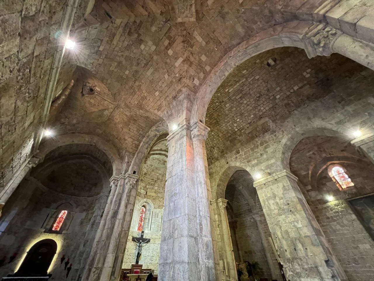 Looking up into the stone vaults of Saint John Mark, columns and arches lit warm, red stained-glass windows