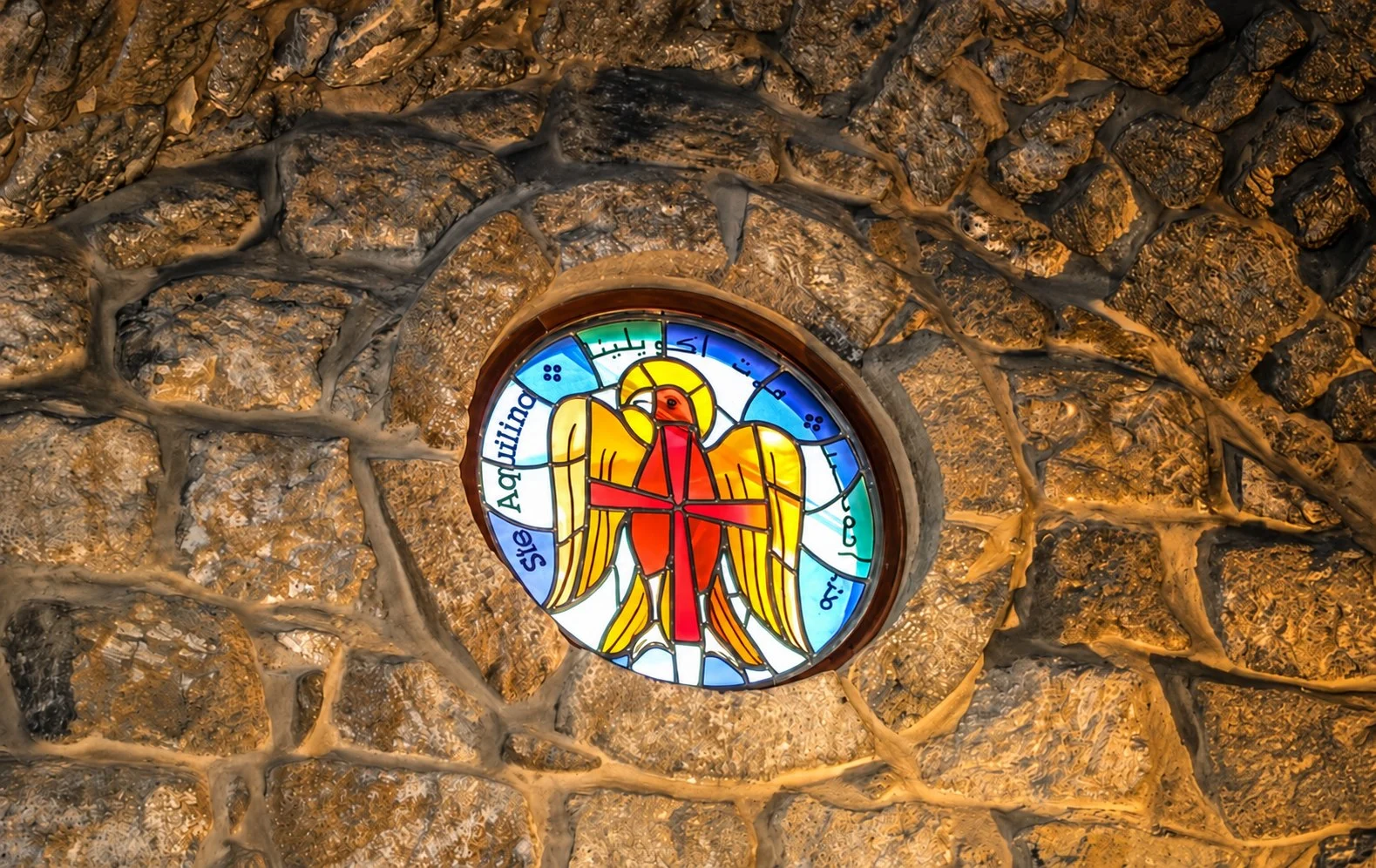 A round stained-glass window of an eagle carrying a red cross, set in the rough stone wall of the shrine