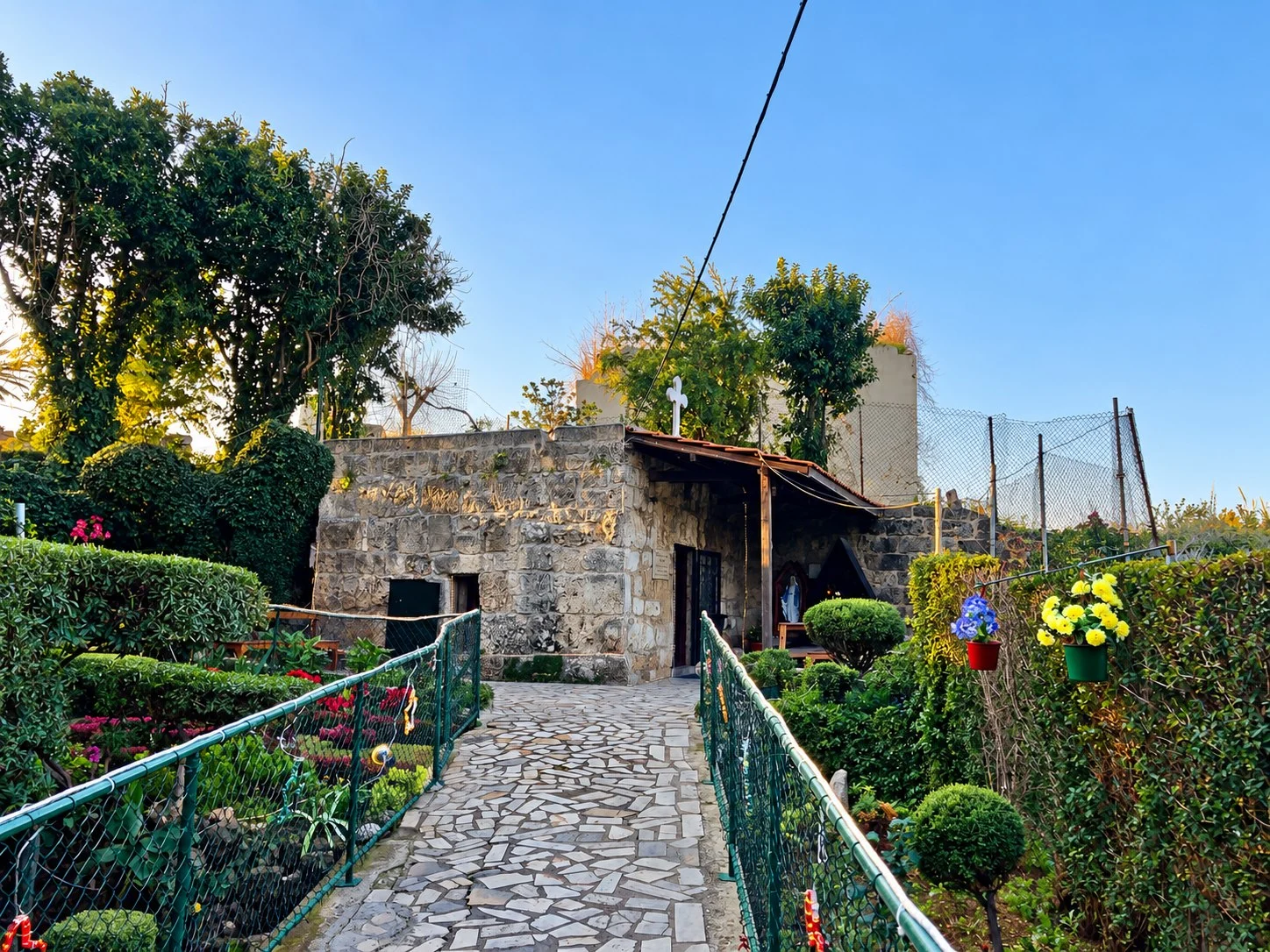 A paved path between green railings and flower beds leading to the low stone chapel of Our Lady of Poverty