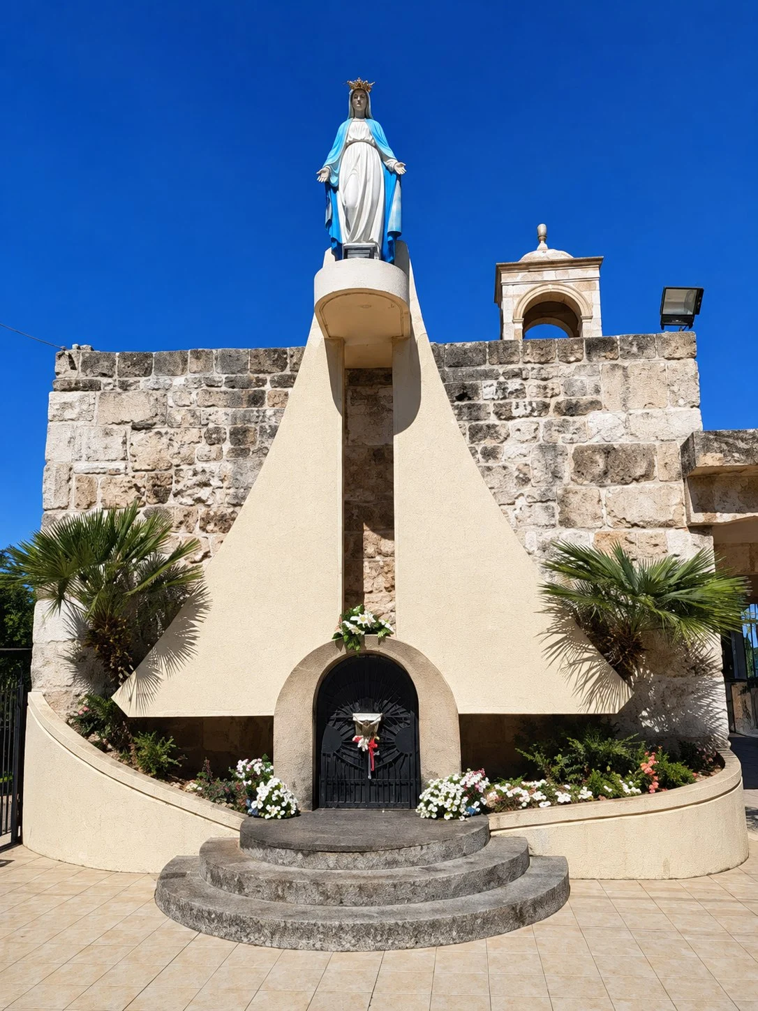 The statue of the Virgin Mary standing high on the white pillar of the shrine, steps and flowers at its foot
